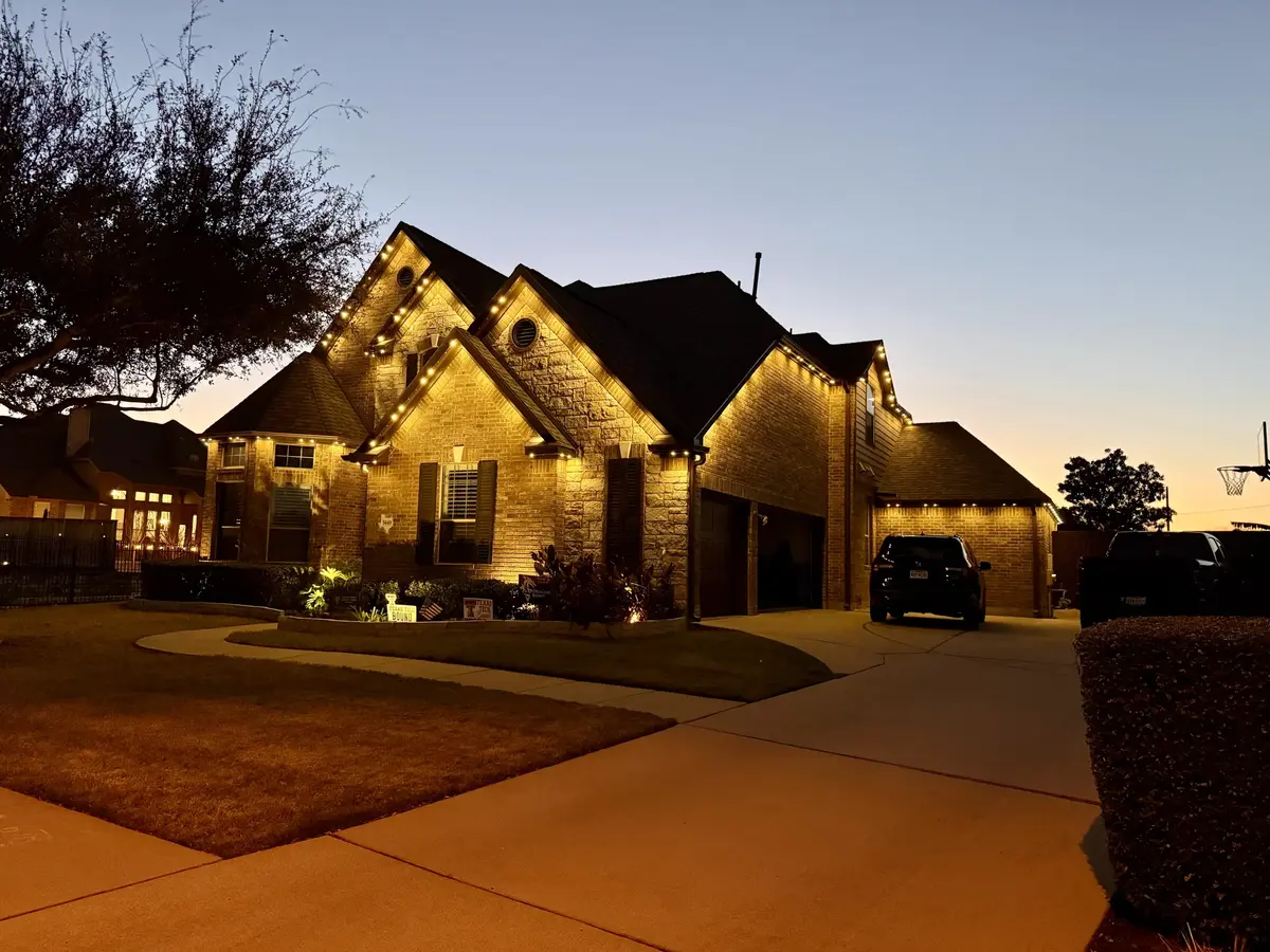 Large brick-and-stone home at dusk with warm white permanent lights along every roofline and gable