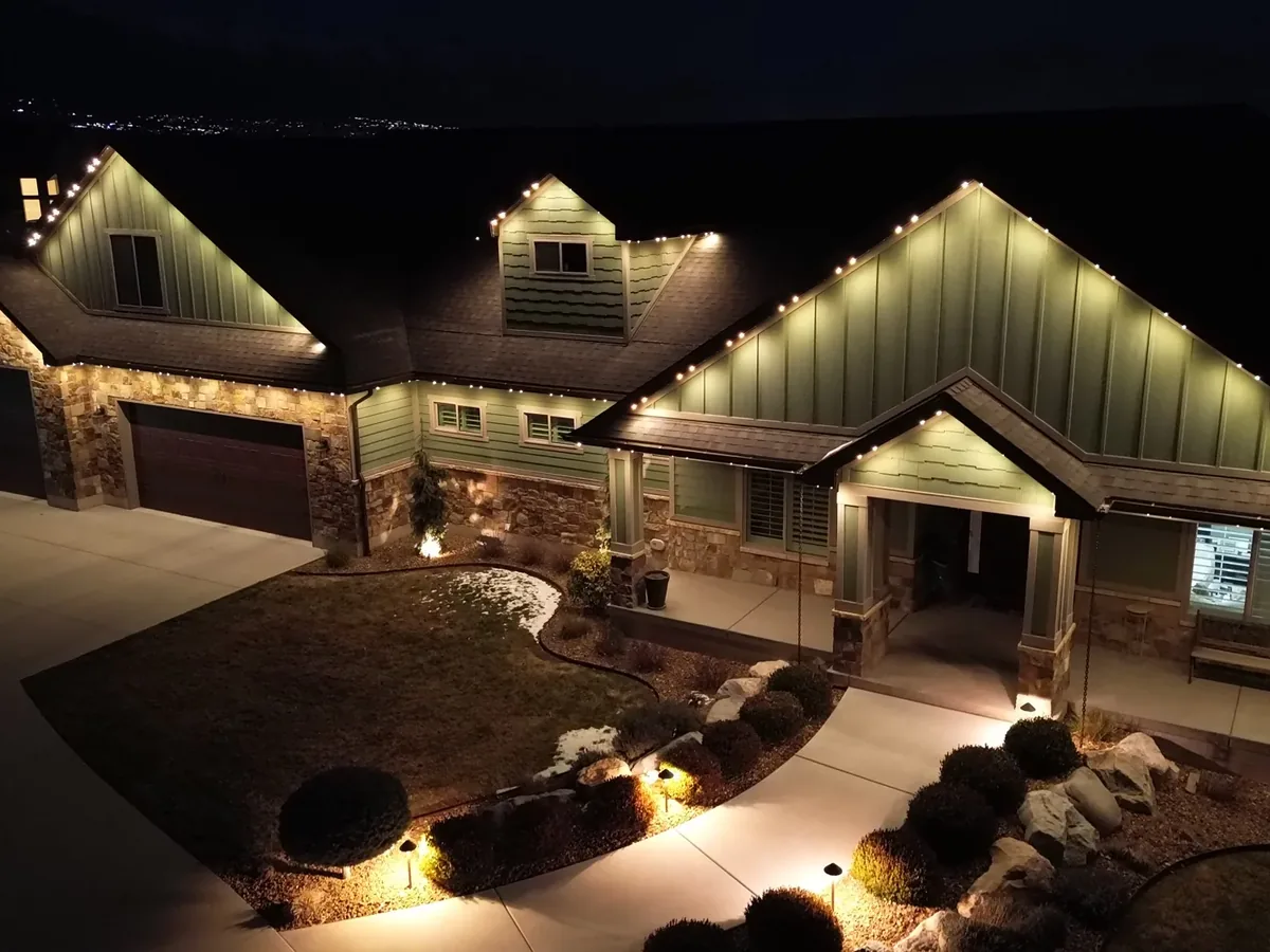 Elevated drone night view of a craftsman home with roofline lights plus lit landscaping and walkway