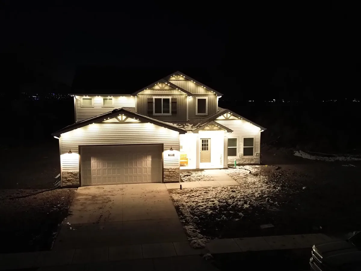 Two-storey light-grey house at night with roofline and eave lights on, seen from a drone