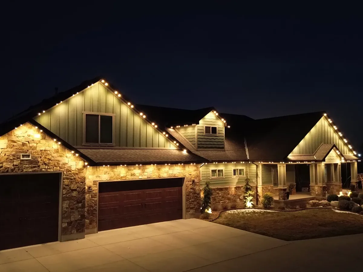 Drone night shot of a stone-and-siding home with warm white permanent lights tracing every roofline and gable