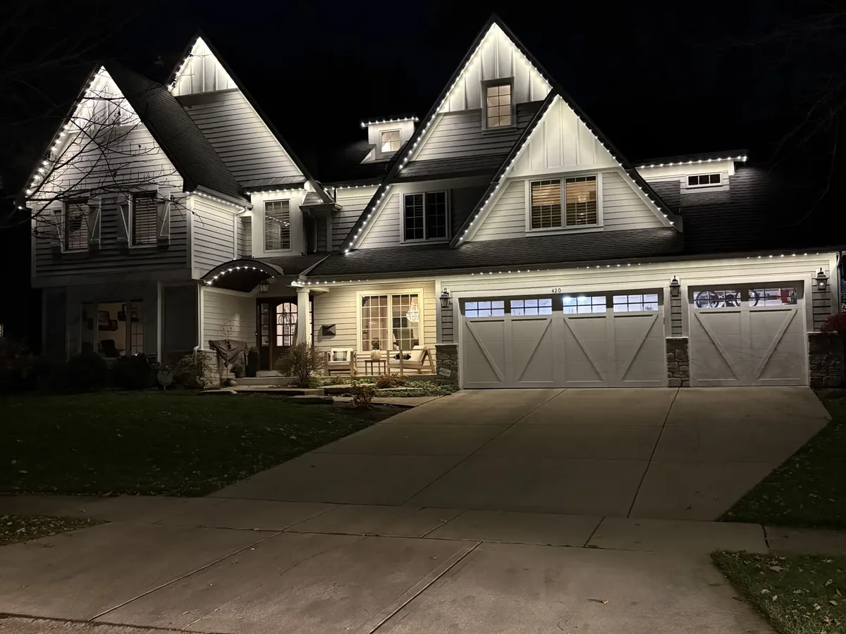 Ground-level night photo of a two-gable house with bright white permanent lights along the gables and garage