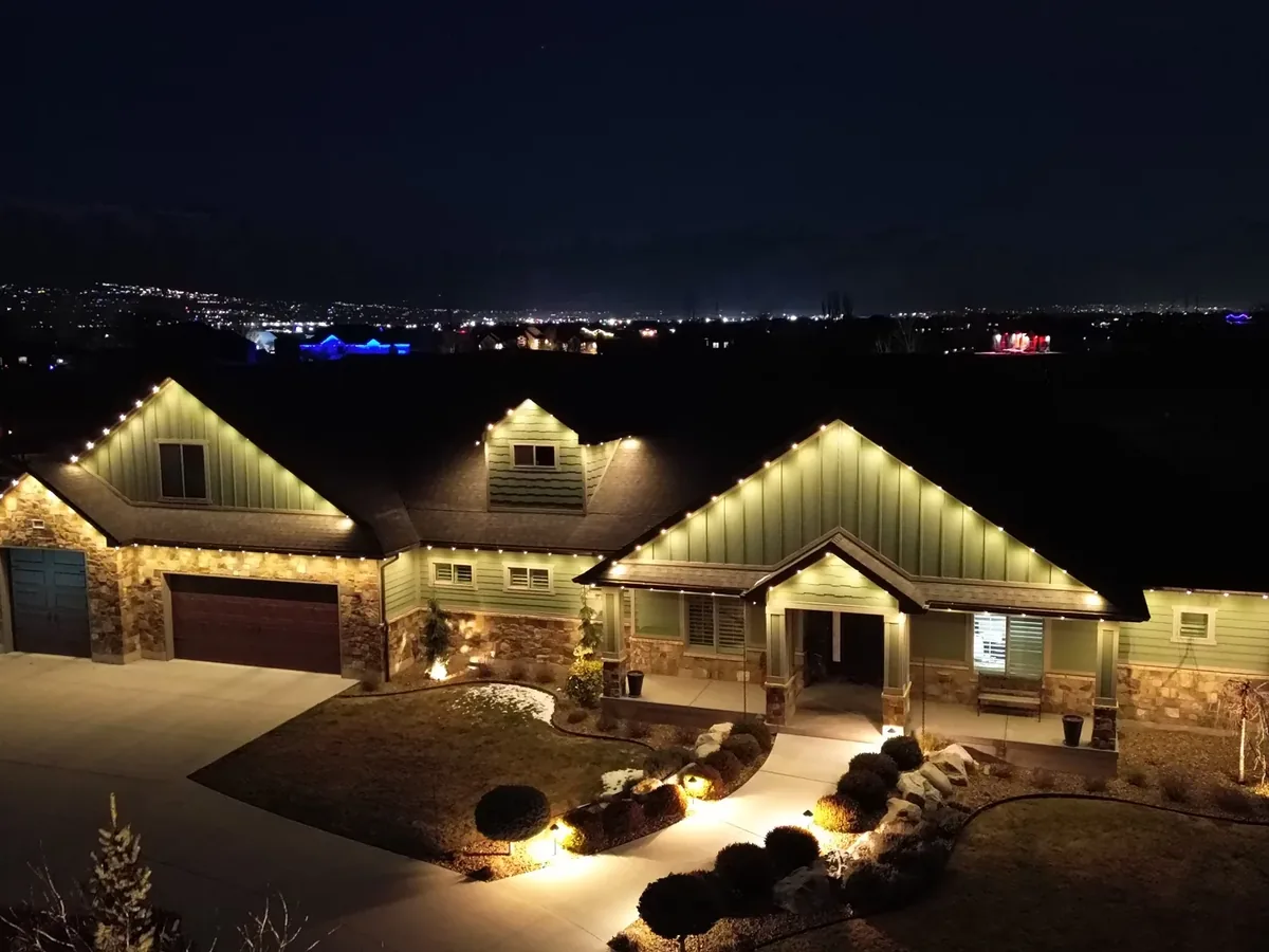 Wide night drone shot of the same style home lit along the rooflines with city lights on the horizon