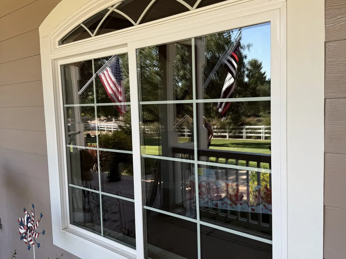 An arched white framed window on a home, clean enough to show the flags and fence reflected in it.