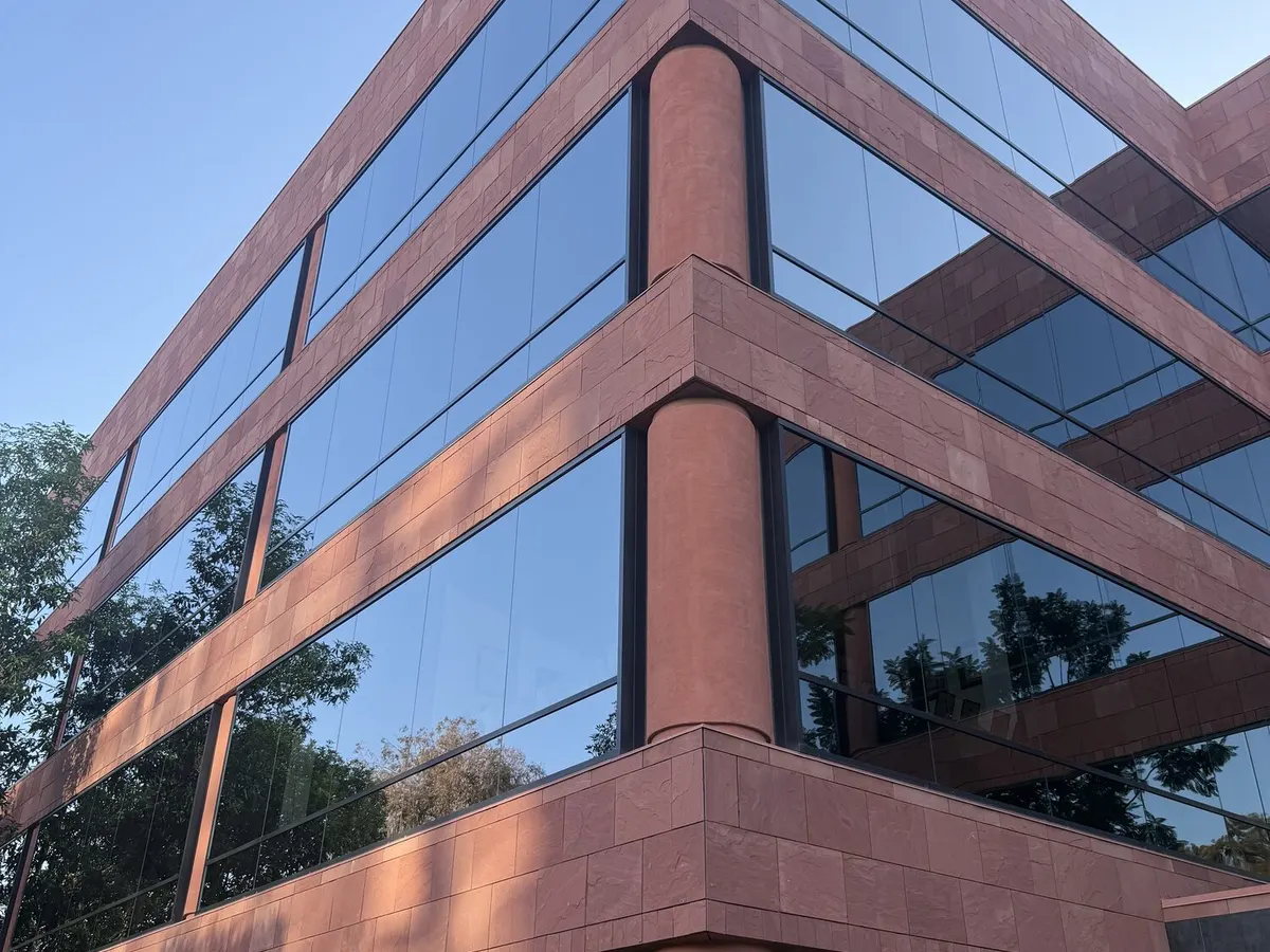 The corner of a four story office building in red stone, its glass clean and reflecting trees and sky.