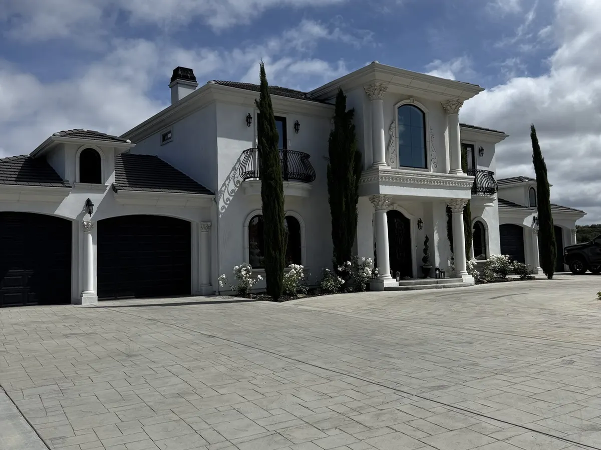 A large white house behind a wide stamped concrete driveway under a cloudy sky.